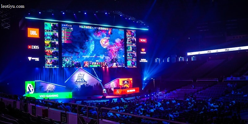 A large esports arena featuring a massive screen displaying a video game, surrounded by sponsor logos and team names. The auditorium is filled with spectators watching the gaming event. Neon blue and purple lights illuminate the stage, creating a vibrant atmosphere.