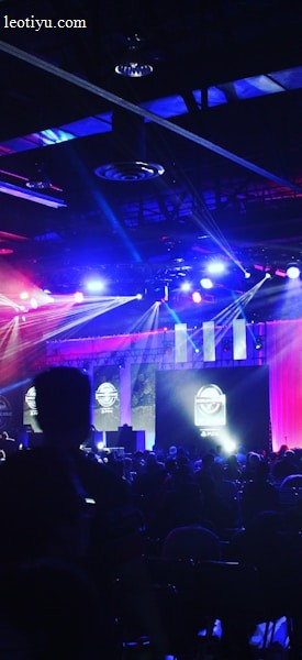 A dimly lit indoor arena filled with an audience. Bright stage lights in red and blue illuminate multiple large screens displaying graphics.