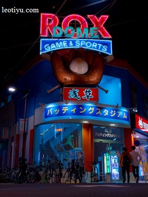 A vibrant, neon-lit facade of a gaming and sports establishment at night, featuring a large glove holding a baseball above the entrance. The building is adorned with colorful signs in both English and Japanese, with various advertisements and an illuminated sign that says 'ROX DOME'. Several people are walking towards the entrance, and bicycles are parked along the side.