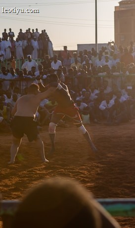 A vibrant wrestling match taking place outdoors, surrounded by a large crowd of spectators. The atmosphere is charged with excitement as people stand and sit around the dirt arena, intently watching the competitors. The sun is setting, casting a warm glow over the scene.