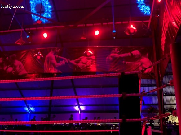 A brightly lit boxing or wrestling ring with vivid red and blue lights. Banners with images of fighters hang from the ceiling. The ropes of the ring are prominently visible, and a stool is placed inside the ring. The arena is filled with an audience, silhouetted against the colorful lighting.